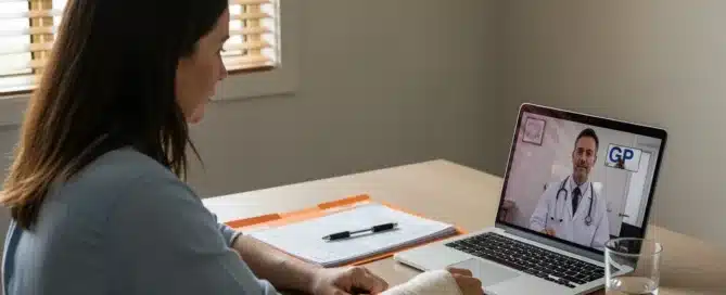 A woman with a bandaged arm sits at a desk having a telehealth video consultation with a male doctor on her laptop.
