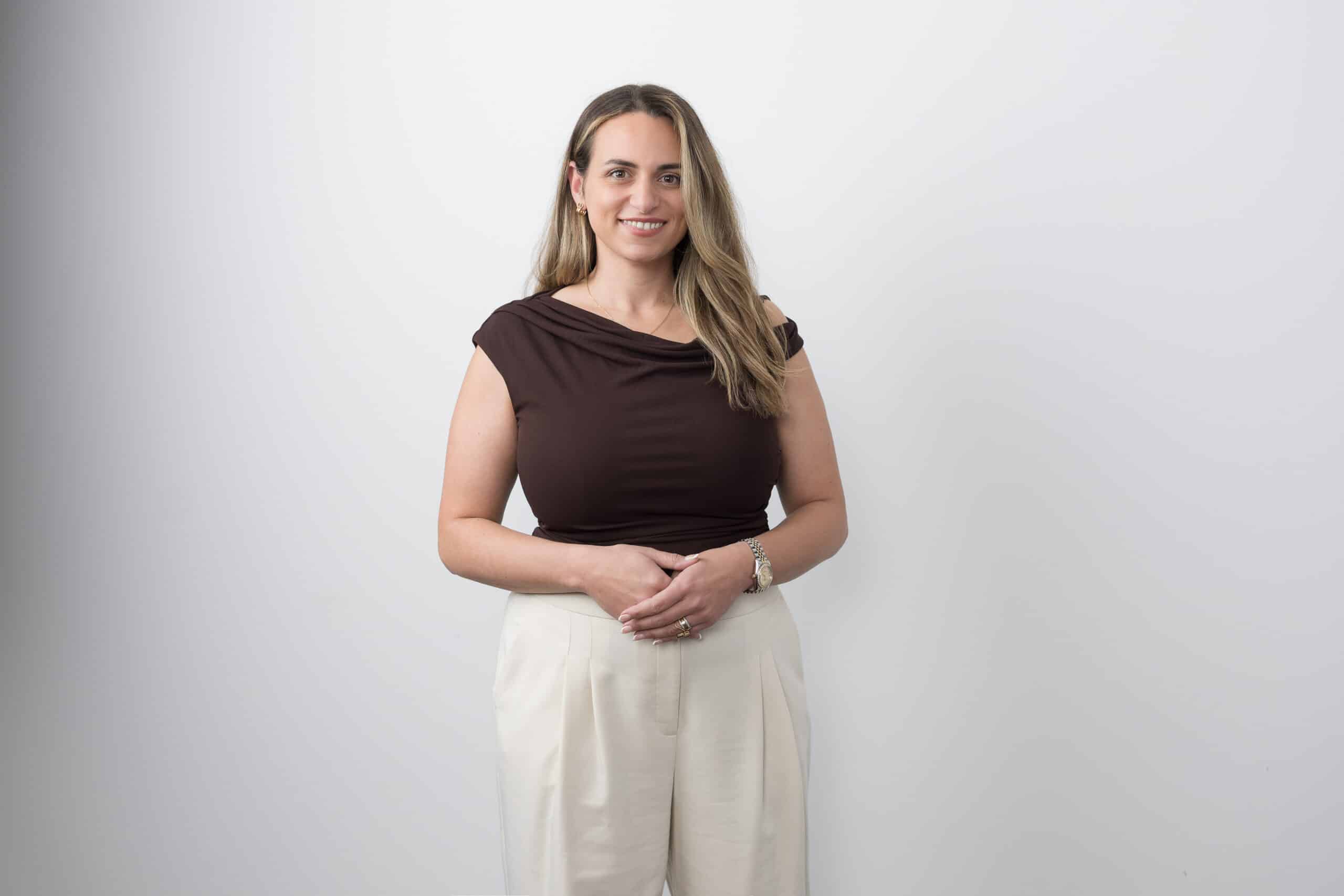 Woman in a brown top and white trousers standing against a pale studio backdrop.