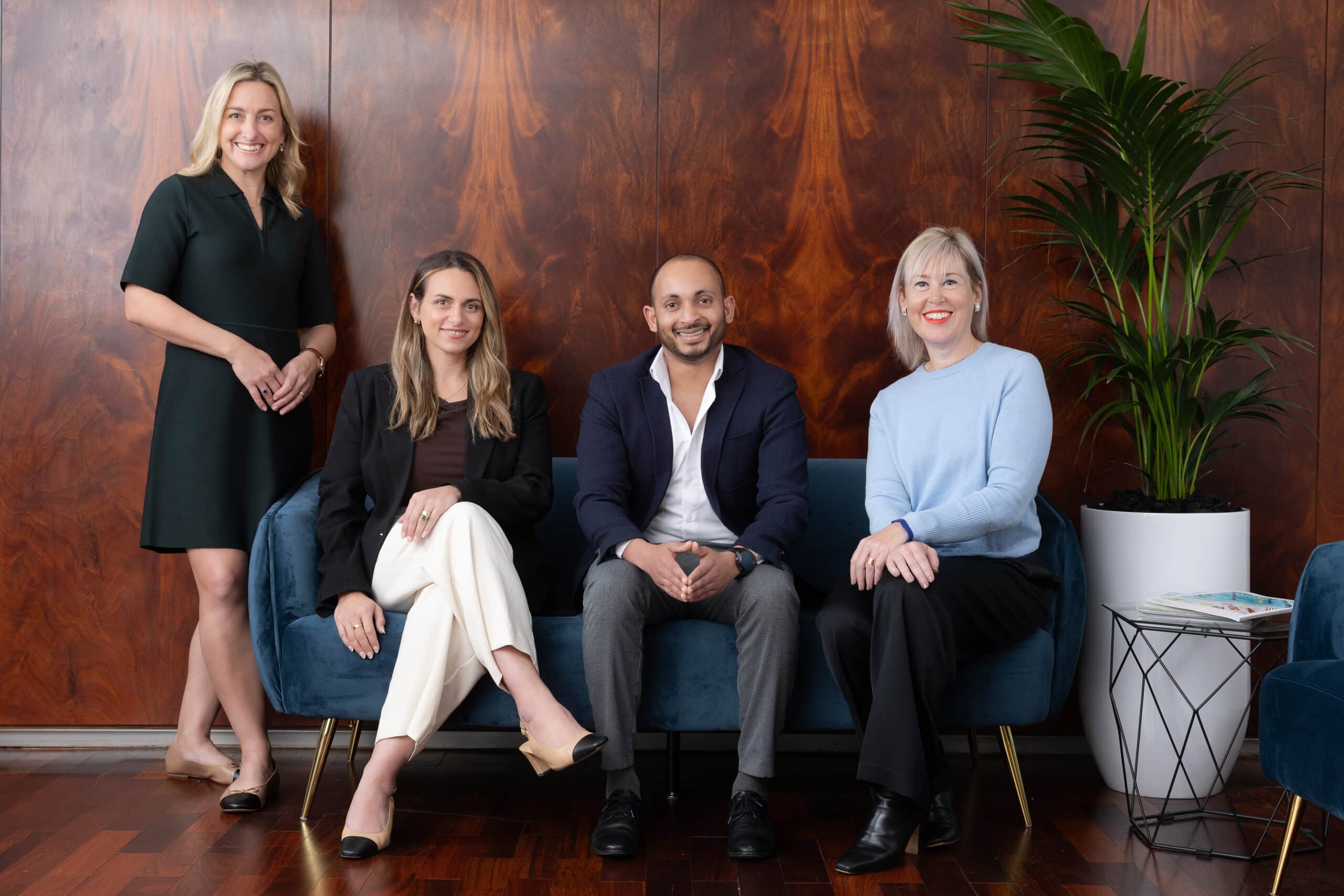 Four professionally dressed people posed in a lounge with a timber wall and large potted plant.