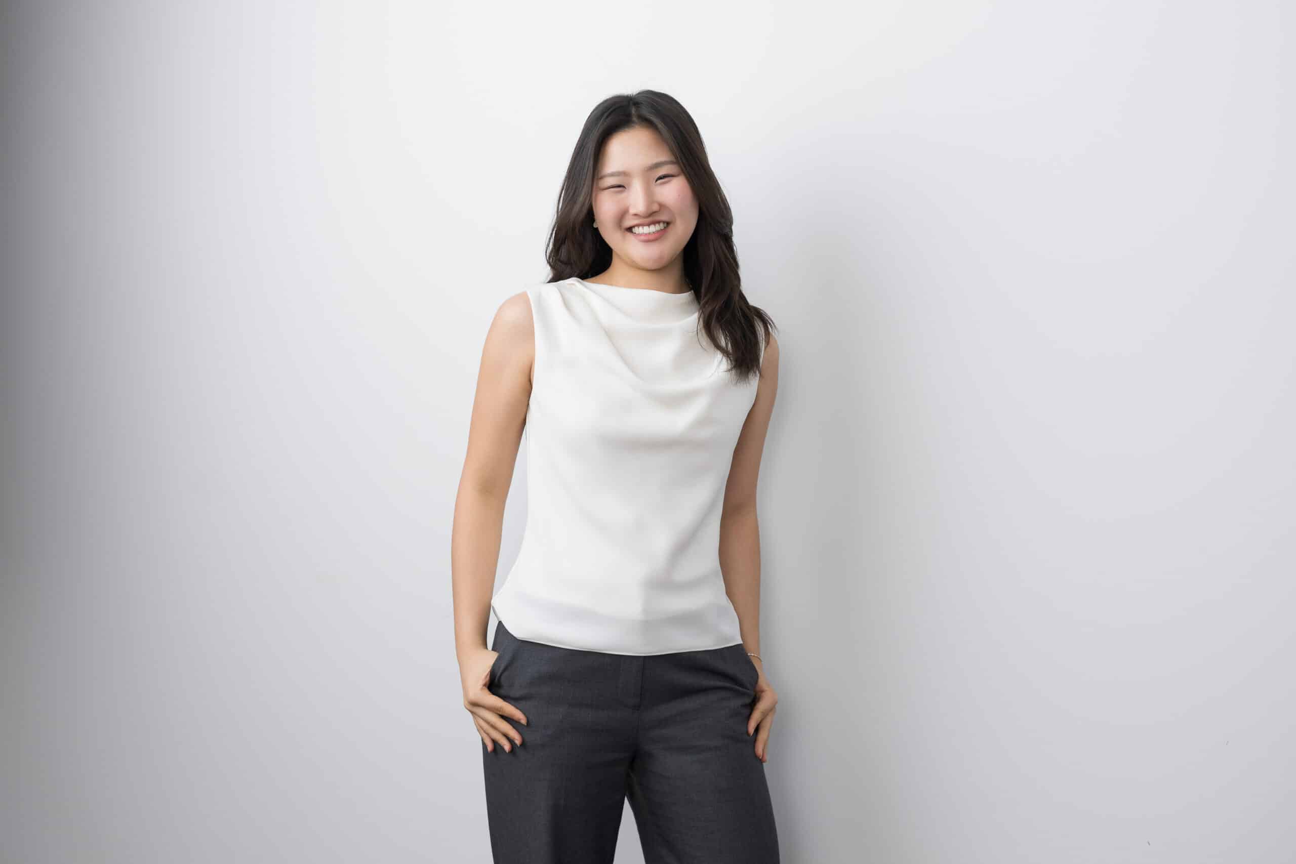 Woman in a white sleeveless top and dark trousers standing against a pale studio backdrop.
