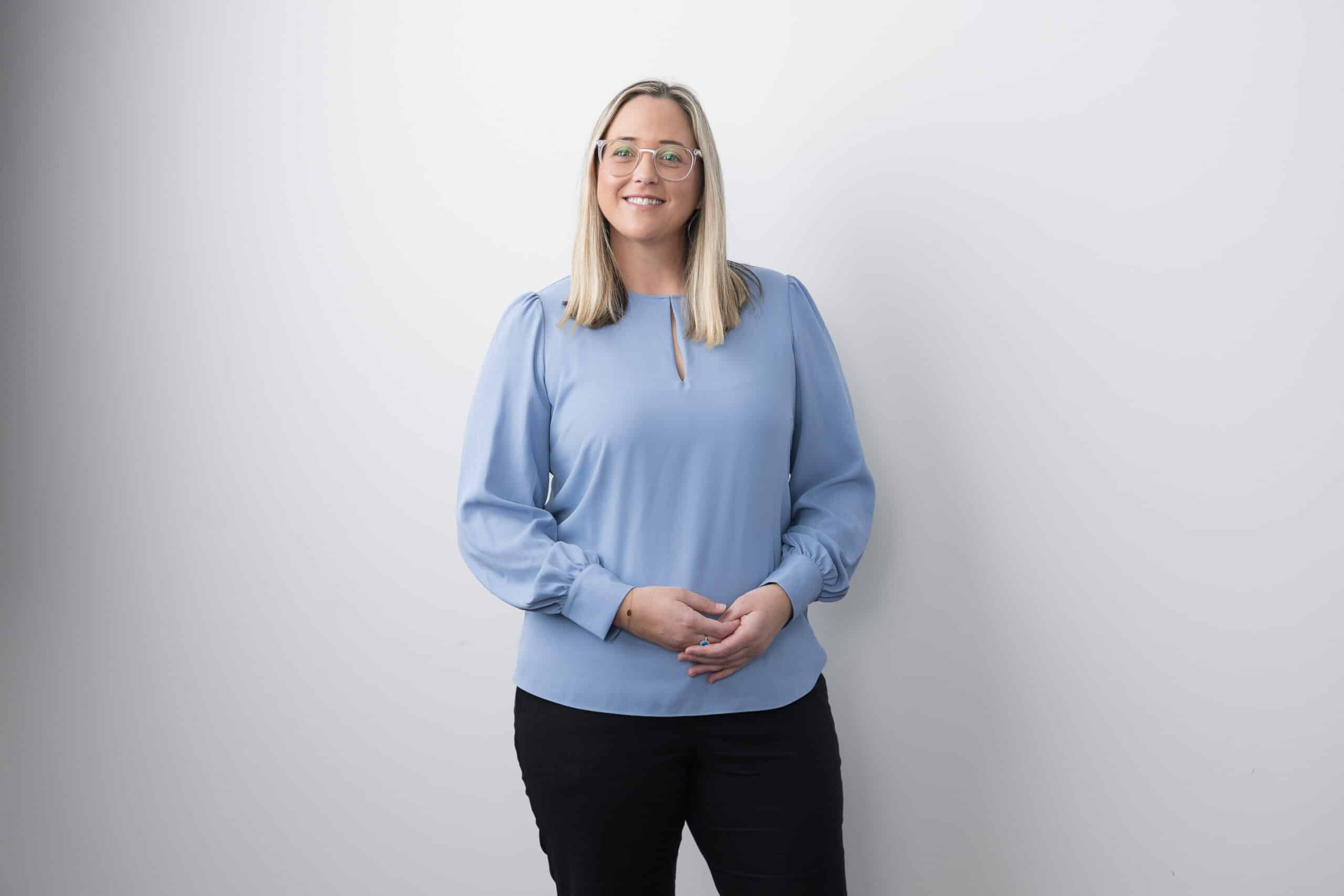 Woman wearing glasses and a light blue blouse standing against a pale studio backdrop.