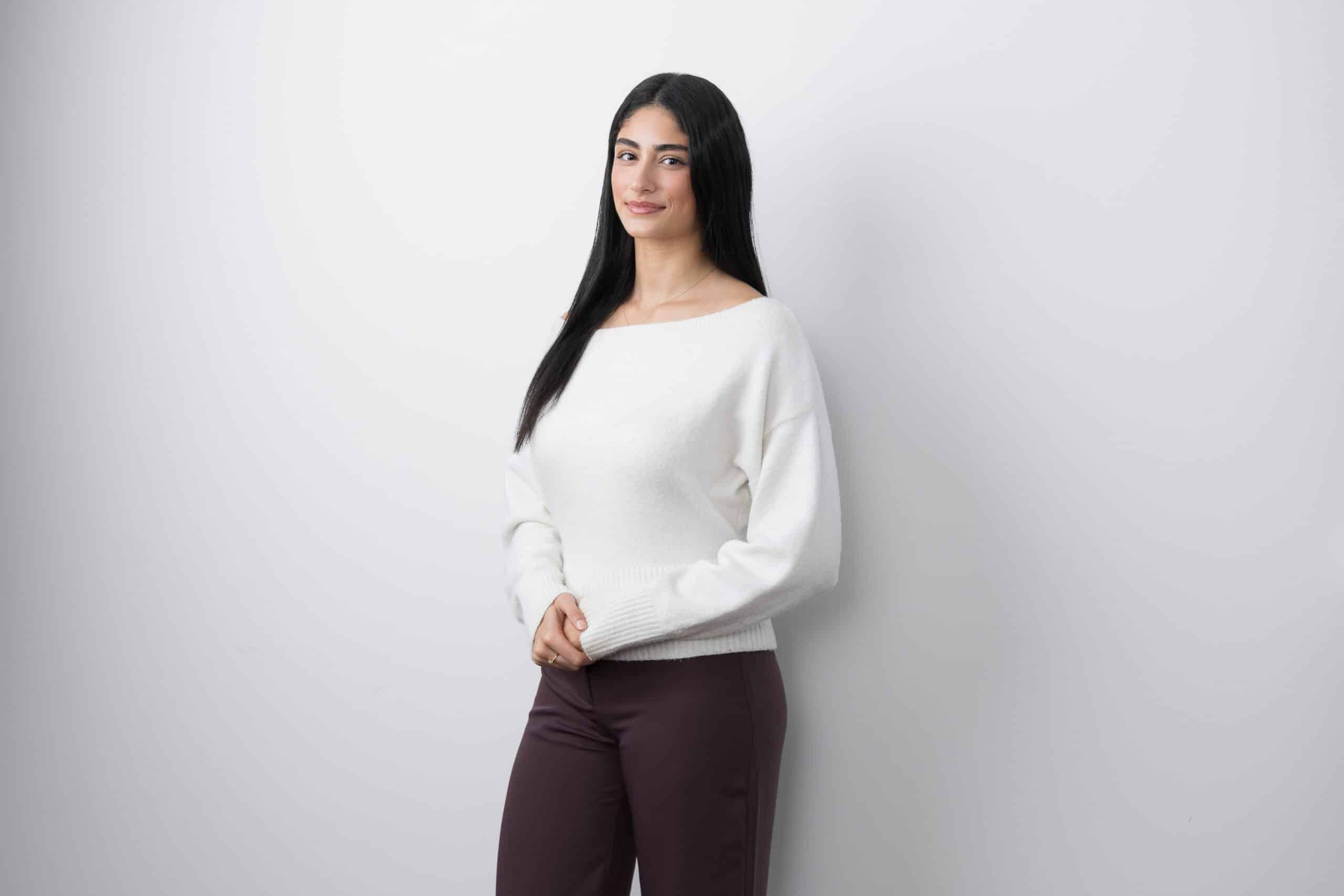 Woman with long dark hair in a white jumper standing against a pale studio backdrop.